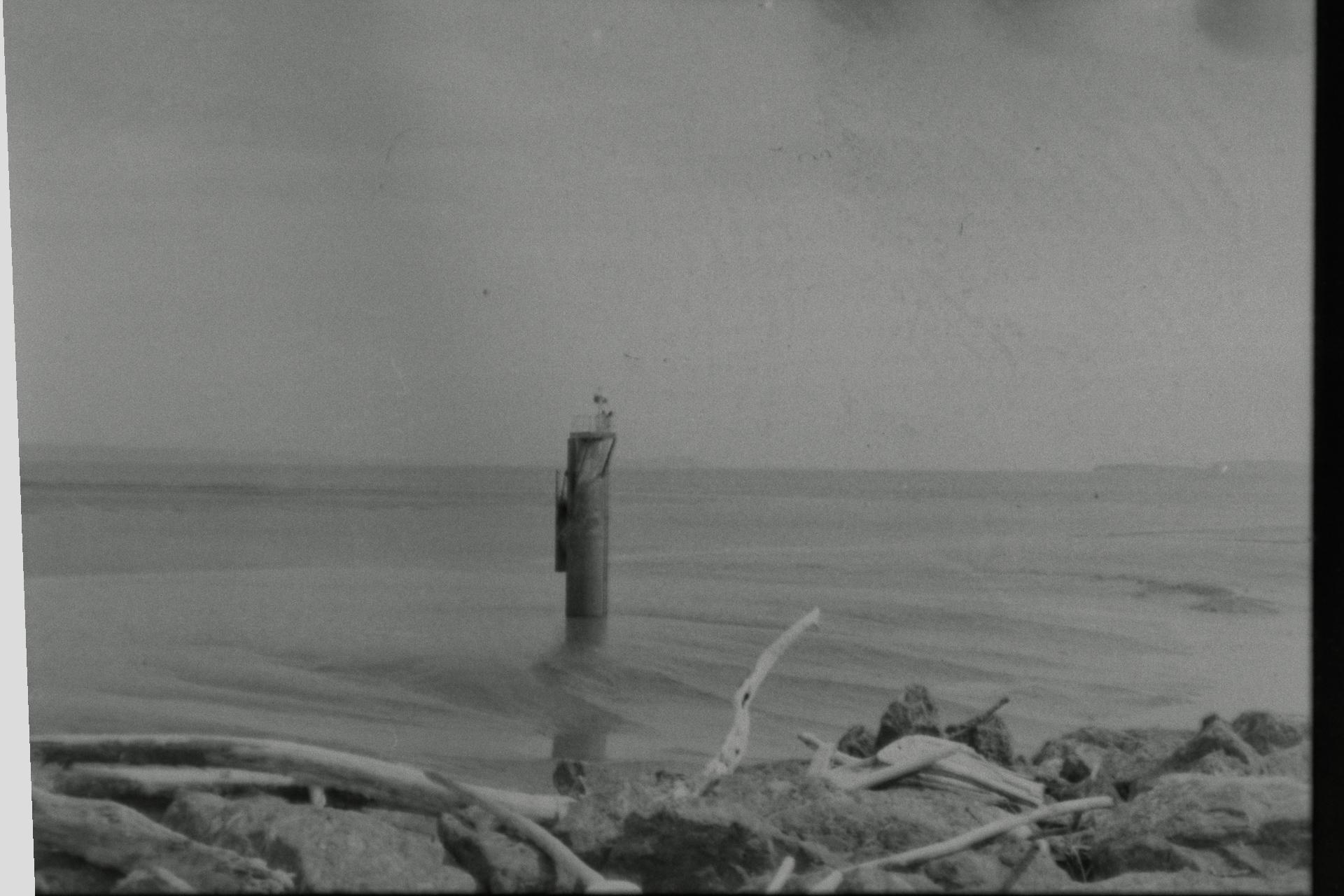 A black and white picture of some kind of big, metal pole with a thing on the front. It's sticking out of mud in the Bristol Channel, with an ethereal white aura around it